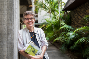 Halbporträt von Prof. Dr. Ingrid Piller an eine Mauer gelehnt mit Buch in der Hand